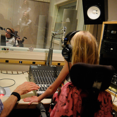 A child facing away from the camera, sitting at an audio mixing board, wearing headphones; an adult hand reaching toward the board from the left.
