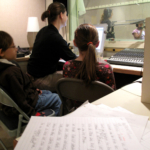 A stack of handwritten papers in the foreground. An engineer and two students at the back in front of a monitor and mixer.