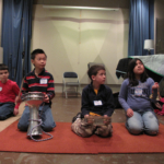 Four students sitting on a carpeted floor, three holding percussive instruments, all looking to the right.