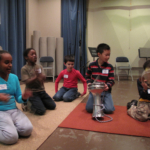 Five students sitting on a carpeted floor, two playing percussions and two clapping, one in the background singing.