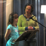 Vocal coach Maria Glanz and a student both wearing headphones, standing in front of a microphone in a recording studio.