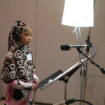 A student in hijab, wearing headphones and standing in front of a microphone and a music stand in a recording studio.