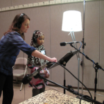 Vocal coach Joy Mills-Parker and a student in hijab, both wearing headphones standing in front of microphones.