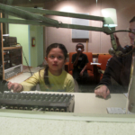 A student turning a knob on a mixer. A woman to her right, looking at her from behind the monitor.