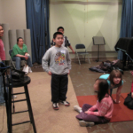 A diverse group of students scattered in a room with a microphone and a piano.