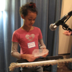 A student popping a piece of bubble wrap in front of a microphone, behind a music stand.