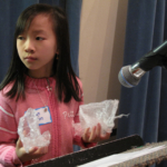 A student holding a bubble wrap piece in each hand in front of a microphone, looking to the left.