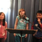 Three students standing in front of a microphone facing the camera, looking to the left.