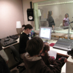 A studio's control room with a woman and three elementary school students. A woman and a student behind the glass window.