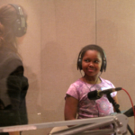 Vocal coach Joy Mills-Parker and a student, both wearing headphones standing in front of a microphone.