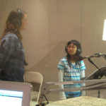 Vocal coach Joy Mills-Parker and a student looking at eachother, both wearing headphones standing in front of a microphone.