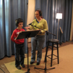 Vocal coach Maria Glanz giving instructions to a student as they both stand in a recording studio in front of a microphone.