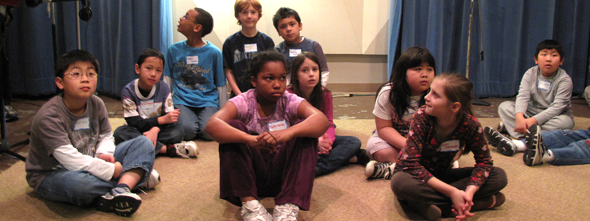 A diverse group of students sitting on a carpeted floor with blue curtains behind them, looking in different directions.