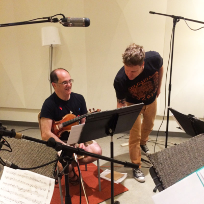 Misha seated with a violin. Peter leans in toward the music stand in front of Misha. Microphones overhead.