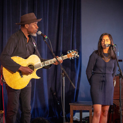 Reggie Garrett, holding an acoustic guitar, and Afrose Fatima Ahmed, on a stage standing at microphones.
