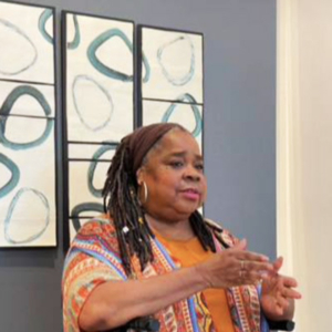 Kathya Alexander speaking in front of an abstract painting of dark circles on a light background.