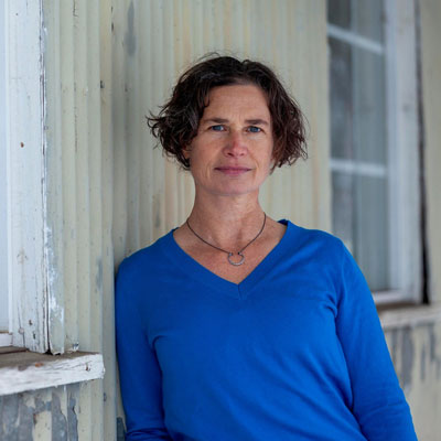 Photo of Perri Lynch Howard in a blue shirt, leaning on a weathered white wall.
