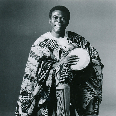 An African-American man is standing in traditional garb, holding a drum under his arms.