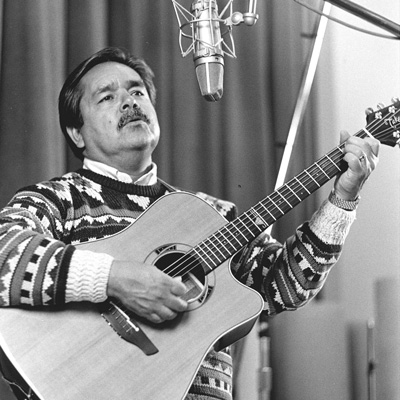 Black and white photo of Juan Barco playing acoustic guitar, standing in front of a microphone.