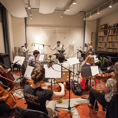 Several musicians in a studio, facing away from the camera. At the back of the room, a saxophonist, conductor, and singer face the camera.