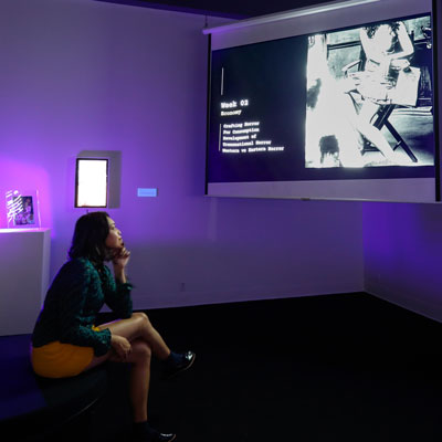 May Maylisa sits on the left in the dimly lit gallery, watching a video projected onto a screen on the right.