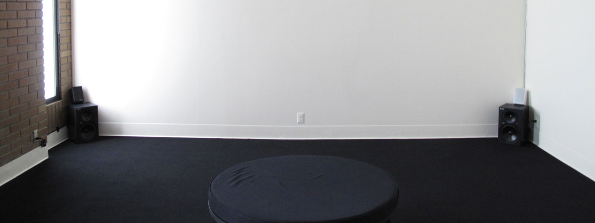 A circular black cushion in the foreground in a room with black carpet, one brick and two white walls. Speakers in the two corners.