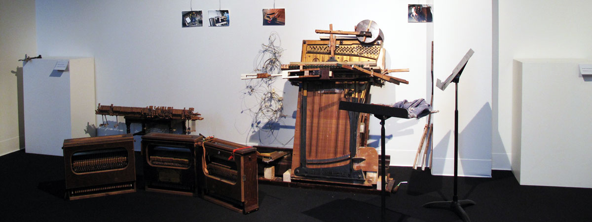 Gallery view, instruments made from the parts of a piano. Black carpet, white walls.