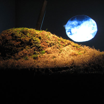 Installation view of F40.298: Generalized Opus Foramina. A mound of mossy earth with a black tube coming out of it; a projected rough circle behind it showing blue sky and clouds.