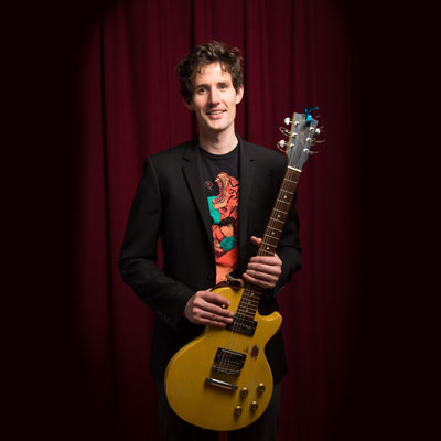 Michael Hamm holding a yellow electric guitar, standing in front of a red curtain