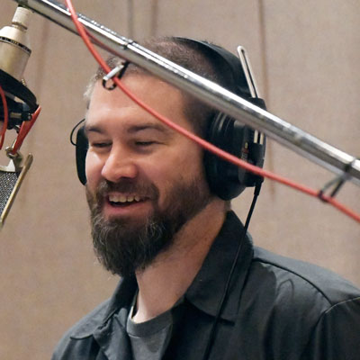 Michael Overa wearing headphones, facing a microphone on the left, smiling
