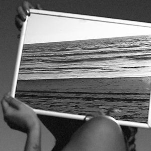 A black and white video still, hands holding a mirror in a white frame, reflecting the ocean and beach