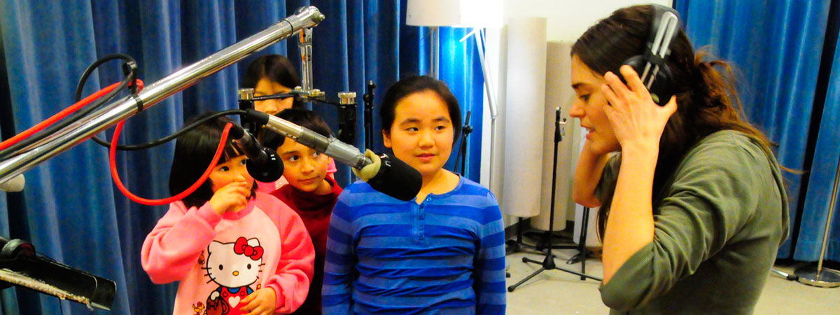 Four students standing behind a pair of microphones watch an audio engineer wearing headphones.