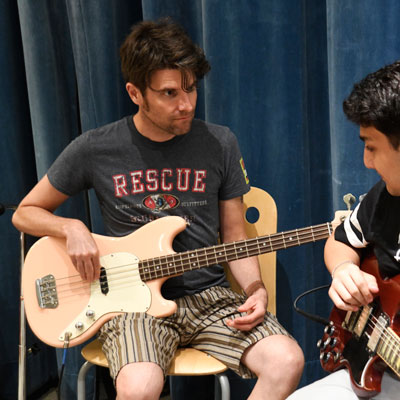 Bill Horist sitting, holding an electric bass, looknig at a student playing electric guitar