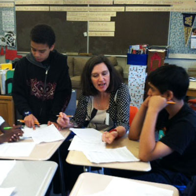 Kathleen Flenniken sits at a classroom table covered with papers, with students on either side of her.