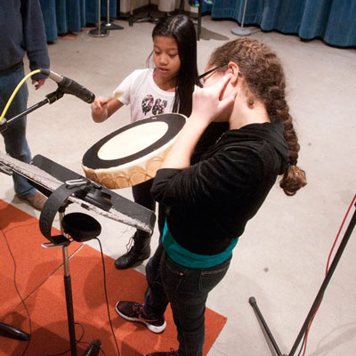 Two students with a frame drum at a microphone. One holds the drum while the other perpares to hit it with a mallet.