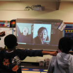 Students watch Sunam Ellis and Brenda Arellano on a projection screen and stretch their arms out.
