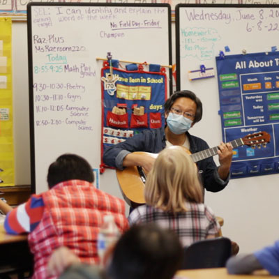 Tomo Nakayama playing guitar in front of several students in a classroom.