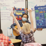Tomo Nakayama holding a guitar in front of several students in a classroom, one holding two thumbs up in the air.