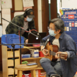Tomo Nakayama sitting with a guitar next to a microphone; Joel Maddox in the background working on a computer.