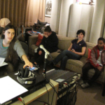 A recording studio control room. On the right, engineer Camelia Jade leans forward with her hand on a knob. on the right, in the background, three people sit on a sofa.
