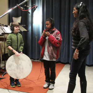 Three students standing, wearing headphones, the one on the right holds a large round drum. Microphones overhead.