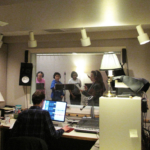 Daniel Guenther faces away from the camera, looking at a compute rmonitor. Through a glass window, Meg McLynn and three students, all wearing headphones.