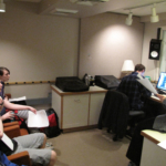 On the left, four students sitting in chairs. On the right, Meg Mclynn faces them holding pages and Daniel Guenther sits with his head down near a computer monitor.