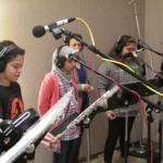 Three students wearing headphones at music stands, with Meg McLynn behind them. Microphones in the foreground.