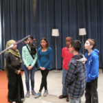 Six students stand around a pair of microphones, lips pursed as if whistling.
