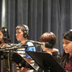 Four students wearing headphones face to the left, with music stands and microphones in front of them.