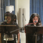 Two students face the camera, wearing headphones, standing at music stands with microphones.