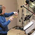 A students wearing headphones stands in front of a microphone and hits a small bell with a pencil.