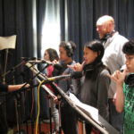 On the left, Camelia Jade adjusts a microphone. On the right, four students face her, holding their hands up with pinky and thumb outspread between their mouths and the microphones. Andrew McGinn stands behind them.