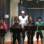 Four students face the camera, all wearing headphones and standing behind music stands with microphones. Andrew McGinn, behind them, also wearing headphones.